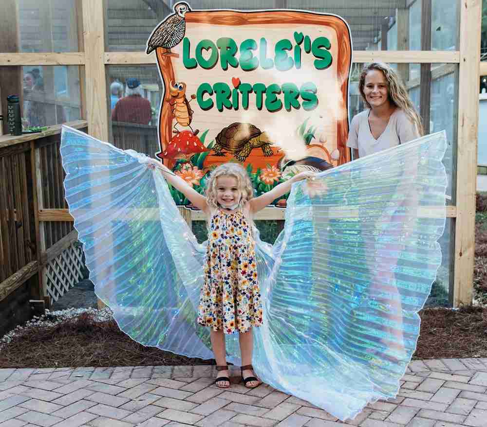 girl wearing butterfly wings mother in background Lorelei's Critters at butterfly garden sign at butterfly garden at boggy creek airboat adventures